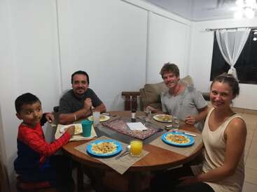 The image depicts a family of four seated around a round wooden table, enjoying a meal together in a cozy dining room with white walls and a window.