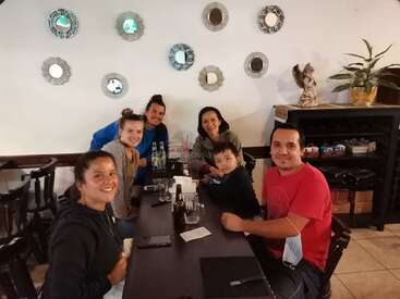 The image depicts a group of six people seated at a long table in a restaurant, surrounded by drinks and a menu, with a white wall adorned with mirrors in the background.