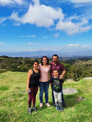 The image depicts a family of four standing together in a lush grassy field, with rolling hills and trees in the background under a blue sky with white clouds.