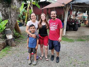 The image depicts a family of four standing together in front of a red barn, with a gravel driveway and lush greenery surrounding them.