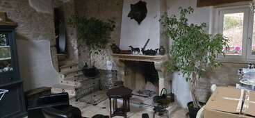 Una acogedora habitación de estilo rústico cuenta con paredes de piedra, una chimenea, árboles en macetas, una escalera, muebles negros, una ventana, cajas de cartón y una decoración ecléctica por todo el espacio.