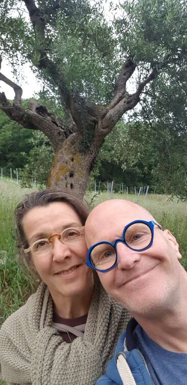 A smiling couple takes a selfie outdoors near an ancient olive tree, surrounded by tall grass and nature. Both wear glasses, enjoying a peaceful day.