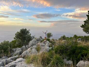 Das Bild zeigt eine Frau, die mit triumphierend erhobenen Armen auf einem Felsvorsprung steht, mit einem ruhigen Gewässer und einem wolkenverhangenen Himmel im Hintergrund.
