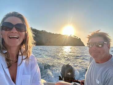 Das Bild zeigt ein Paar mit Sonnenbrille und weißer Kleidung auf einem Boot im Meer, hinter dem die Sonne untergeht, und eine kleine Insel im Hintergrund.