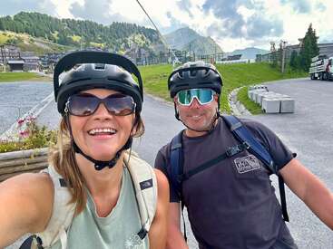 The image shows a selfie of a man and woman wearing helmets and backpacks, standing on a road with mountains and buildings in the background, likely taken during a bike ride.