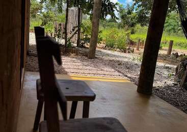 Two rustic wooden chairs sit on a shaded porch overlooking a gravel yard, trees, and garden, with sunlight filtering through branches and a blue sky above.