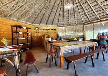 This cozy rustic kitchen features wooden benches, tables, and shelves, with a thatched roof, brick walls, decorative cloths, and a few people dining together.