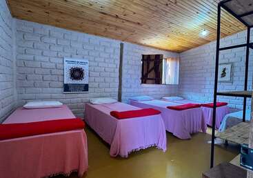 The image shows a cozy room with four single beds covered in pink blankets. The beds have red throws, white pillows, and rustic brick walls.