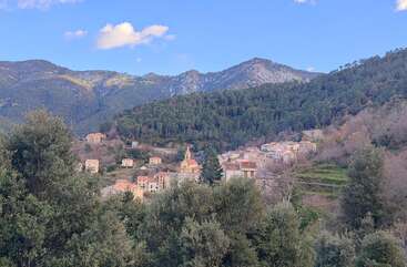 Ein malerisches Bergdorf, eingebettet in üppiges Grün, mit charmanten Häusern, einer Kirche und sanften Hügeln unter einem klaren blauen Himmel mit vereinzelten weißen Wolken.