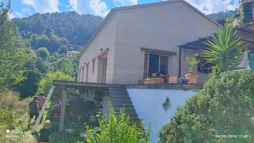 Ein beiges Haus mit einer Terrasse und einem Garten ist von üppigem Grün und Bergen umgeben. Mehrere Menschen spazieren unter einem strahlend blauen Himmel durch den Garten.