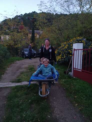 Eine Frau schiebt zwei lächelnde Kinder in einer Schubkarre einen ländlichen Weg entlang, umgeben von Grün, gelben Blumen und Menschen im Hintergrund in der Abenddämmerung.