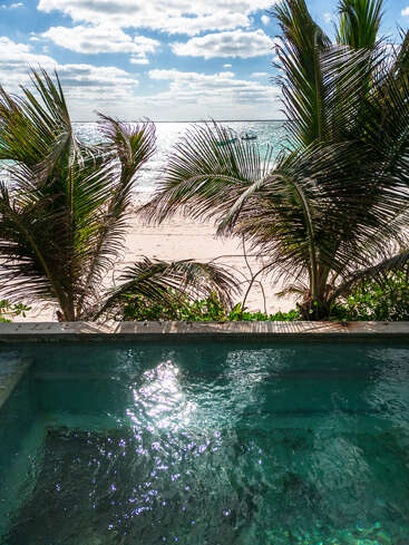 Ein glitzernder türkisfarbener Pool blickt auf einen Sandstrand, der von üppigen Palmen gesäumt ist. Das Sonnenlicht tanzt auf dem Wasser, dahinter liegen das Meer und der blaue Himmel.