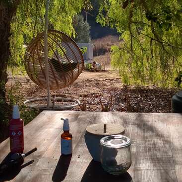 Sunlight filters through green leaves onto a wooden table with bottles and jars. In the background, a hanging rattan chair and nature create a peaceful scene.