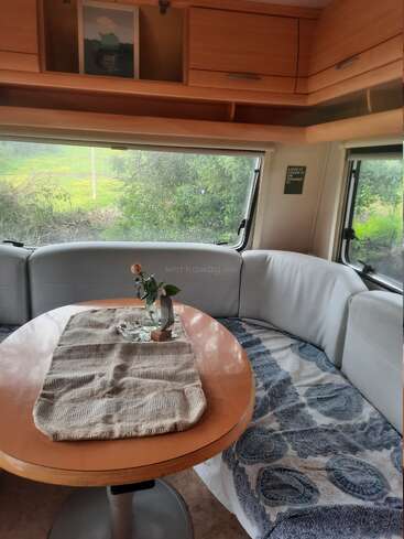 Un acogedor rincón comedor en una caravana con asientos acolchados, una mesa redonda de madera y un sencillo centro de mesa. Los grandes ventanales dejan ver la exuberante vegetación del exterior, creando un ambiente tranquilo.