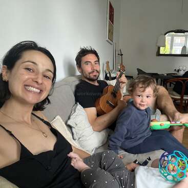 A happy family sits together on a couch; the woman smiles, the man plays a ukulele, and two young children play with toys, creating a cozy atmosphere.