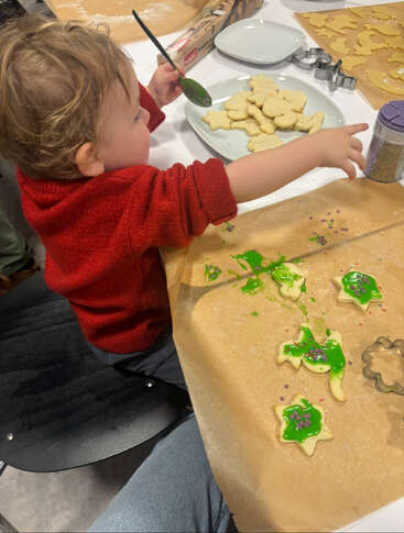 A young child in a red sweater decorates cookies with green icing and sprinkles, sitting at a table filled with cookie cutters, dough, and baking supplies.