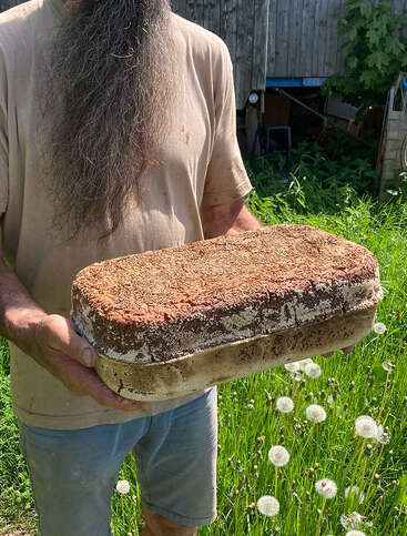 Um homem de barba comprida, vestindo uma camisa bege, segura um grande tijolo retangular coberto de matéria orgânica, em pé num jardim verdejante.
