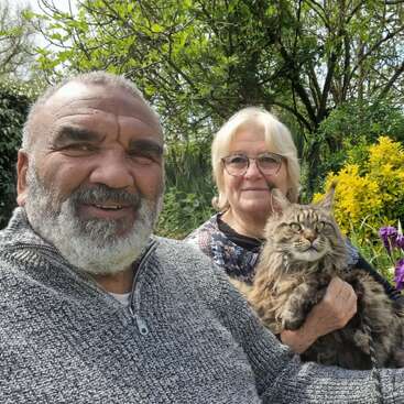 Ein älterer Mann und eine ältere Frau sitzen draußen im Garten und lächeln in die Kamera. Die Frau hält eine große, flauschige Katze im Arm. Um sie herum stehen Bäume und blühen Blumen.