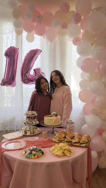 Deux filles sourient lors d'une fête d'anniversaire sur le thème du rose, avec un ballon en forme de « 14 », entourées de ballons, d'un gâteau, de mini-burgers, de bonbons, de chips et de desserts sur la table.