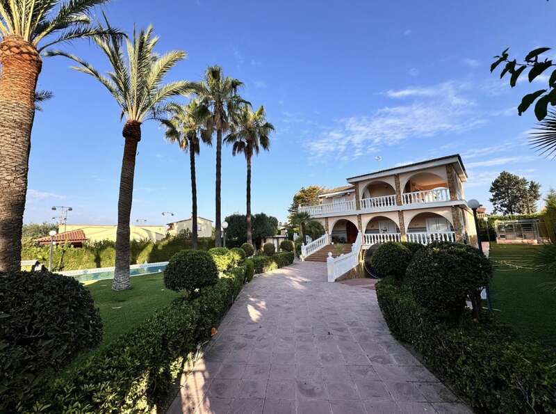 Une magnifique villa à deux étages avec des balcons voûtés se dresse au milieu de jardins soigneusement entretenus, de palmiers et d'arbustes taillés, sous un ciel bleu limpide, créant une atmosphère sereine.