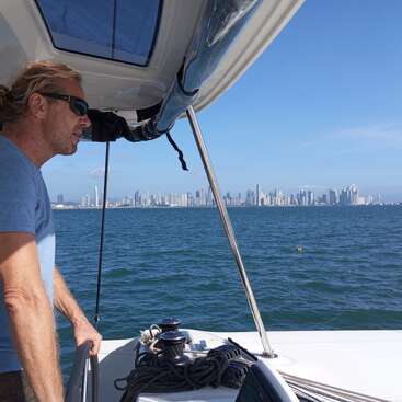 Das Bild zeigt einen Mann, der auf einem Boot steht und auf das Wasser hinausschaut, während in der Ferne die Skyline einer Stadt unter einem klaren blauen Himmel zu sehen ist.