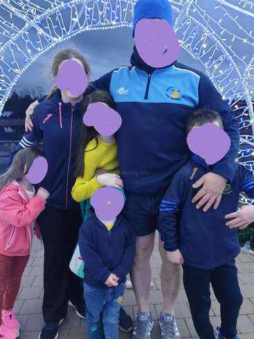 The image shows a family of six posing for a photo, with the father standing in the middle and the children surrounding him, all wearing casual clothing.