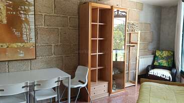 Cette image montre une pièce chaleureuse avec des murs en pierre, une table blanche et des chaises, des étagères en bois, un miroir, un fauteuil avec un coussin et la lumière du soleil.