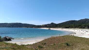 Une magnifique scène de plage avec une eau d'un bleu limpide, du sable blanc, de douces vagues, des dunes verdoyantes, des collines au loin, et des gens qui se détendent et profitent de cet environnement ensoleillé et paisible.