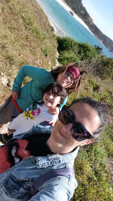 Trois personnes, deux enfants et un adulte, sourient pour un selfie sur un sentier côtier. En arrière-plan, on aperçoit l'eau bleue, la plage de sable et les collines verdoyantes.