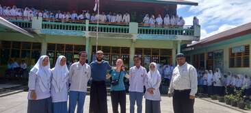 A group of students and teachers pose together outside a school building. Many students are gathered on the balcony and in the courtyard, smiling under a sunny sky.