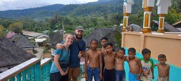 A group of children and two adults pose together on a balcony. Thatched-roof houses, lush greenery, and mountains are visible in the scenic background.
