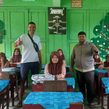 A classroom painted green, with students sitting at blue-desk tables. Two adults stand smiling and giving thumbs up. Educational posters and tree murals decorate the walls.