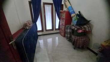 A small, tidy room with three tall windows covered by blue curtains. There are stacked plastic drawers, folded blankets, a rolled-up mat, and a mattress.