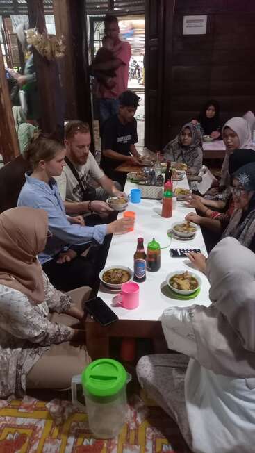 Un grupo de personas, entre las que hay extranjeros y lugareños, se sientan alrededor de una mesa baja, disfrutando juntos de la comida tradicional en un acogedor restaurante de ambiente cálido y comunitario.