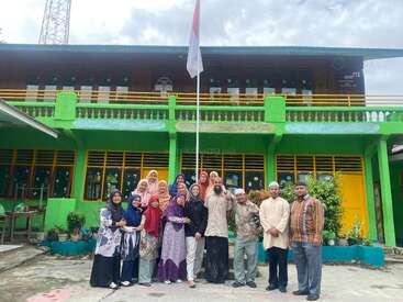 Eine Gruppe von Menschen steht an einem bewölkten Tag lächelnd vor einem grünen Schulgebäude unter einem hohen Fahnenmast, an dem eine indonesische Flagge weht.