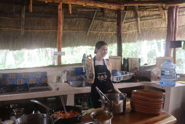 A woman stands in a rustic, open-air kitchen with a thatched roof. She wears an apron, gestures "okay," surrounded by pots, dishes, and cooking ingredients.