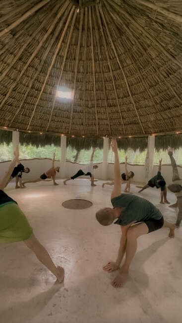 A group of people practice yoga together in a circular, open-air hut with a thatched roof, natural light, and a peaceful, serene atmosphere.