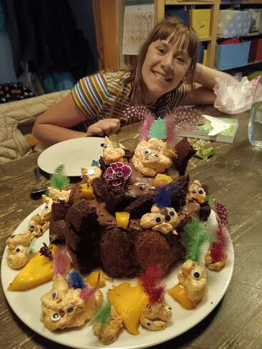 Dans une pièce chaleureuse, une jeune fille souriante est assise à une table devant un gâteau au chocolat fantaisiste, décoré de créatures colorées à plumes, d'yeux mobiles et de tranches de mangue.