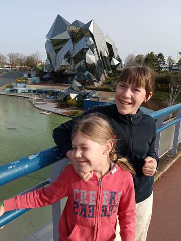 Deux enfants souriants se tiennent sur un pont, profitant d'une journée ensoleillée. Derrière eux se dresse un bâtiment futuriste aux façades miroitantes, à l'architecture angulaire unique, entouré d'eau.