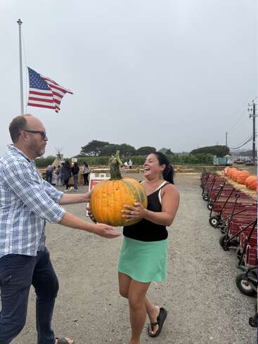 A man and woman joyfully share a large pumpkin at a pumpkin patch. American flag waves in background, with red wagons and people nearby.