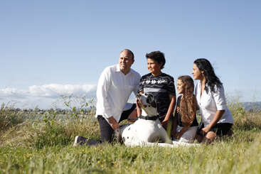 Das Bild zeigt eine vierköpfige Familie, darunter zwei Kinder und eine Frau, die an einem sonnigen Tag mit einem großen weißen Hund in einem Feld mit hohem Gras posiert.