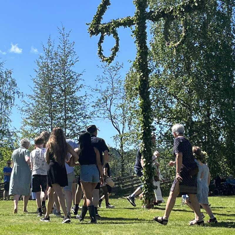 A imagem mostra um grupo de pessoas reunidas em torno de um mastro, adornado com folhas verdes, tendo como pano de fundo árvores e um céu azul claro em um dia ensolarado.