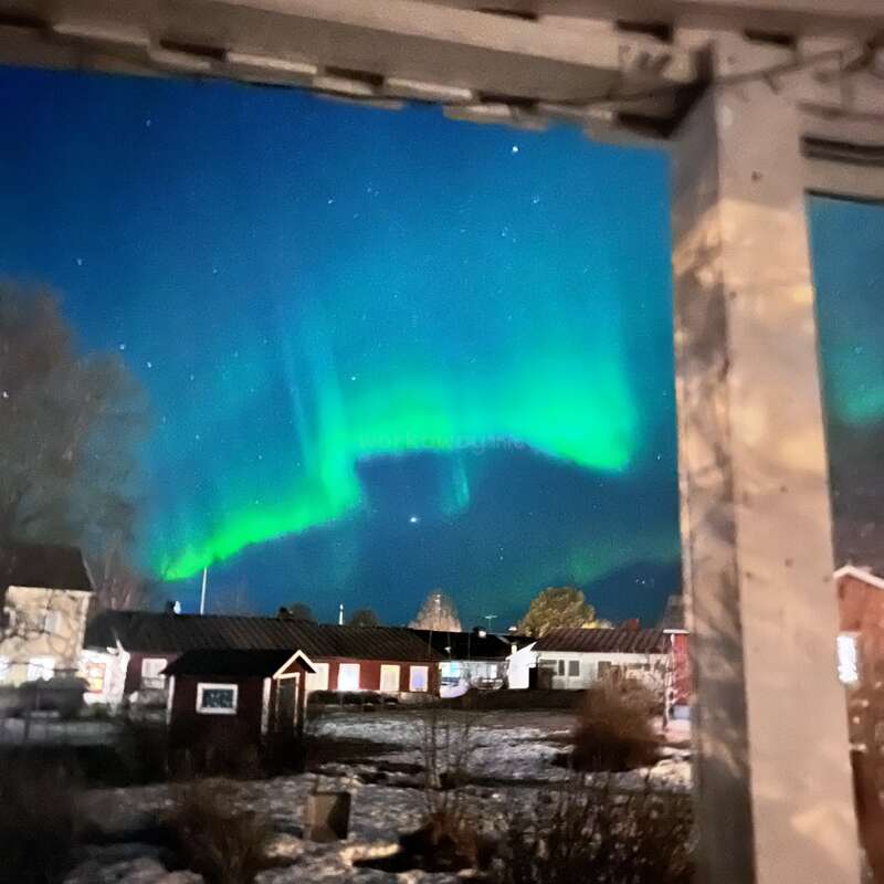 Das Bild zeigt eine ruhige Winterszene mit einem leuchtend grünen Polarlicht am Himmel, vor dem Hintergrund von Häusern und einem schneebedeckten Boden.