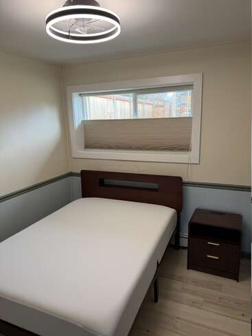 This image shows a small, simple bedroom with a modern ceiling light, single bed, nightstand, window with blinds, and light-colored walls with a green accent.