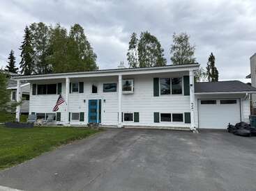 A white two-story house with teal door and shutters, front porch, American flag, single-car garage, driveway, green lawn, and tall trees in the background.