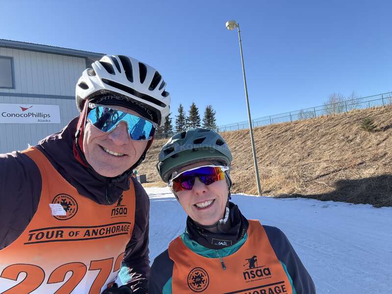 Two people wearing helmets and sunglasses smile outdoors. They are dressed in orange bibs labeled "Tour of Anchorage." Snow, blue sky, and a building are visible.