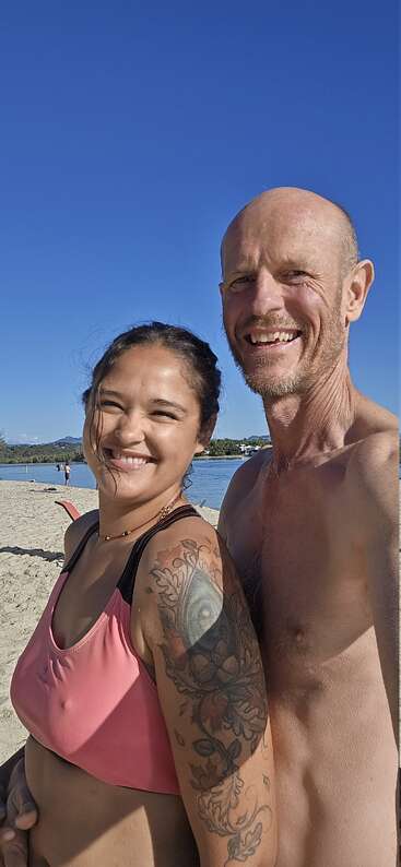 Um casal sorridente está próximo em uma praia ensolarada. A mulher tem uma tatuagem floral no braço; ambos parecem felizes, apreciando o céu azul e a água.