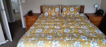 A neatly made bed with a yellow and white floral bedspread sits between two wooden nightstands, each holding a lamp and other small items. Doorway visible.