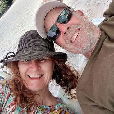 A smiling man and woman are lying on a sandy beach, wearing hats and sunglasses, enjoying a sunny day together, capturing a happy, relaxed selfie moment.