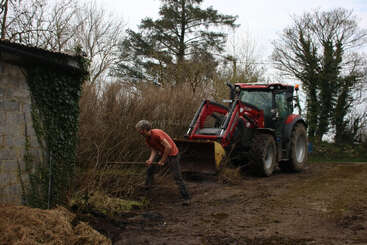 A person is working outside, using a pitchfork to clear debris near bushes and a wall, with a red tractor parked behind them on a muddy path.